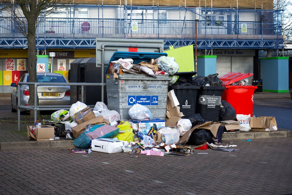 A collection of black garbage bags and discarded cardboard boxes stacked against the base of a red metal door embedded in a textured, off-white exterior wall. The bags are piled haphazardly, some lean
