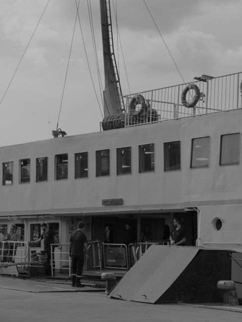 A small passenger ferry with a blue hull and white upper structure navigating through a river or harbor during late afternoon or early evening, with sunlight casting a glow on the scene. The ferry has